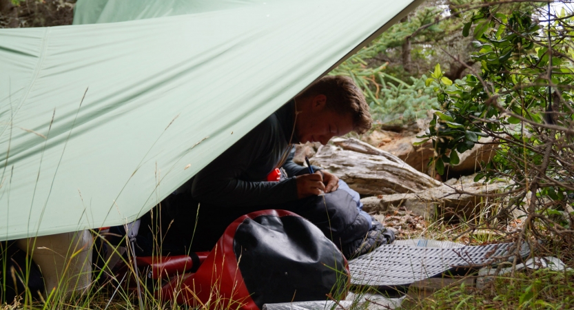 From under a tarp shelter, a person journals. 